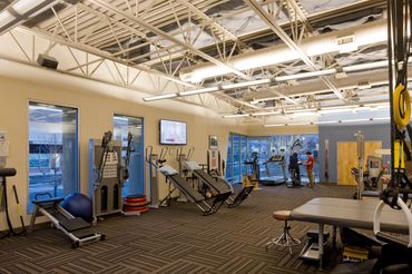 Spacious physical therapy gym with exercise equipment, open ceiling structure, and natural light.
