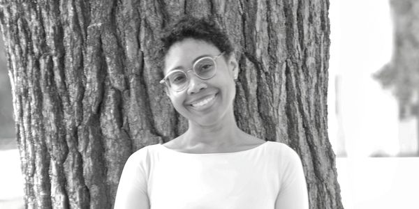 Smiling person with glasses leaning against a tree.