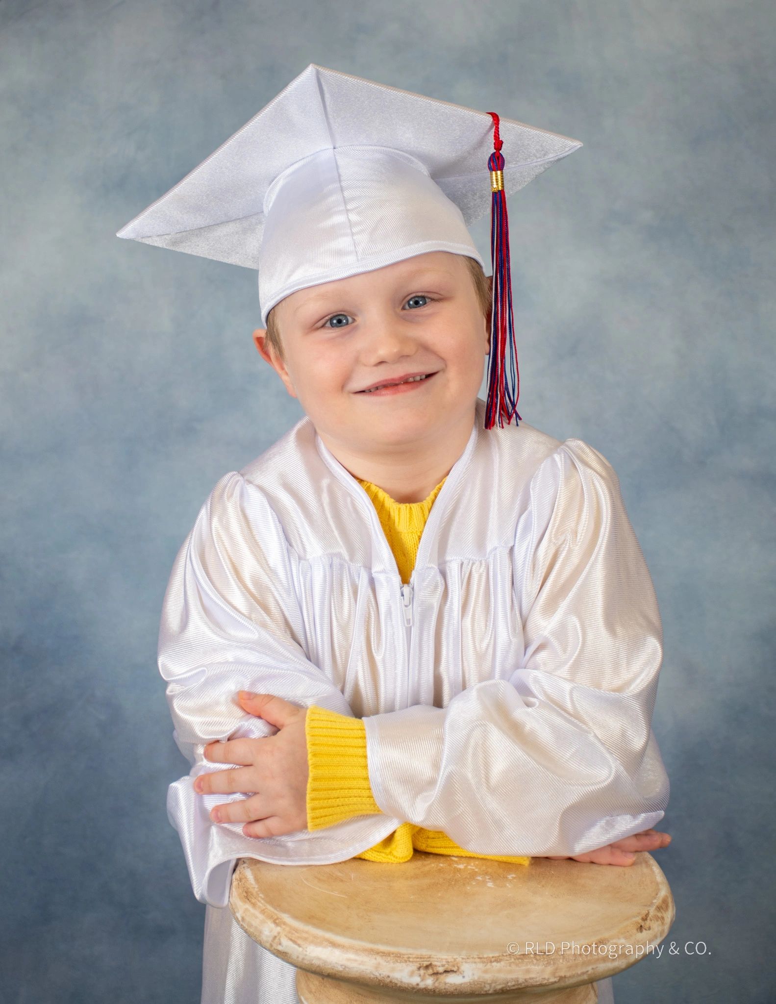 Young boy wearing a white graduation cap and gown, with arms folded, leaning on a podium smiling.