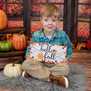 A boy holding a "hello fall" sign surrounded by pumpkins and fall decor.