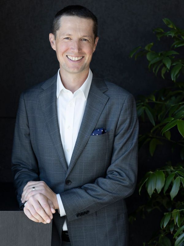 Smiling man in a gray suit with a white shirt, standing beside a plant.