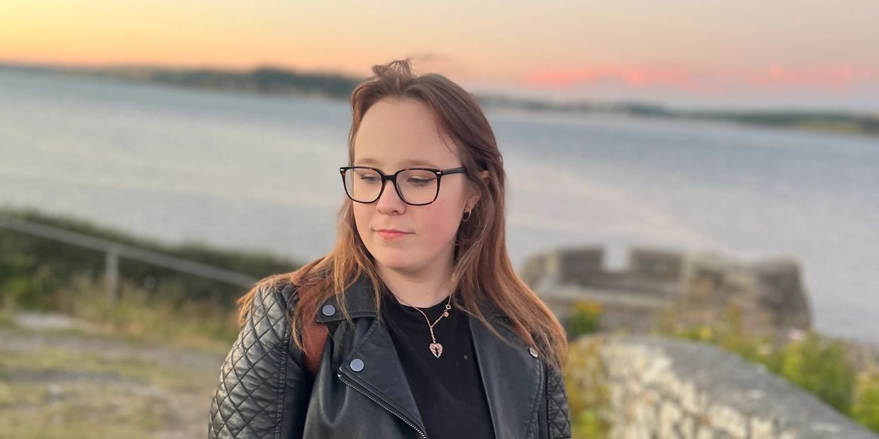 A young woman with glasses in a black leather jacket by the water at sunset.