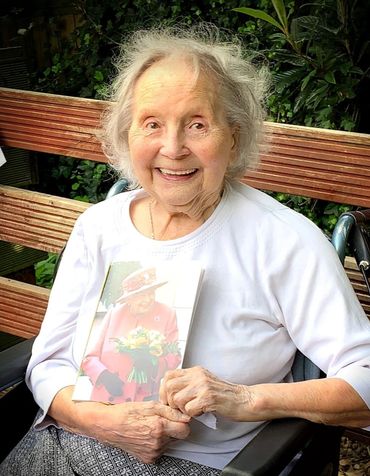 Elderly woman in a wheelchair smiling and holding a card, celebrating her 100th birthday.
