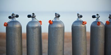 A row of six scuba diving air tanks standing on the beach.