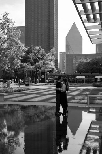 A couple embraces in a city park with reflective water and tall buildings.