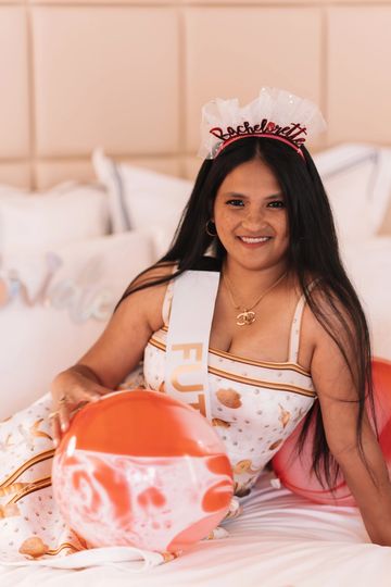 A woman wearing a bachelorette sash and headband sits on a bed holding an orange balloon.
