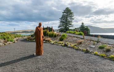 Wooden sculptures line a gravel path with scenic hills and water in the background.