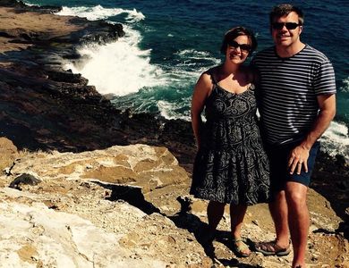 Couple smiling on rocky coast with ocean waves behind them.