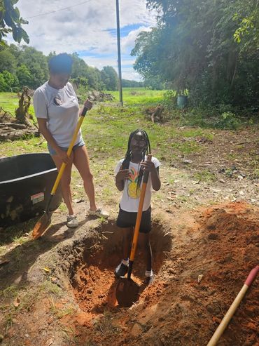 Two girls digging a hole with shovels in a sunny outdoor area.
