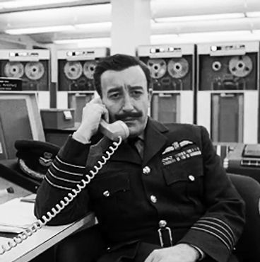 Man in military uniform talking on a phone in a vintage control room.