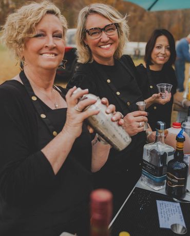 Three women bartenders smiling while making drinks outdoors.