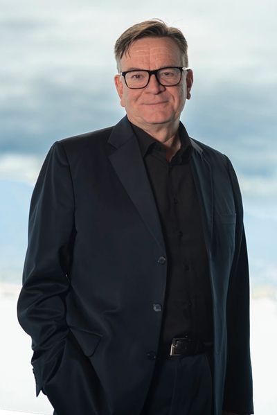 Man in a black suit and glasses posing confidently with hands in pockets.