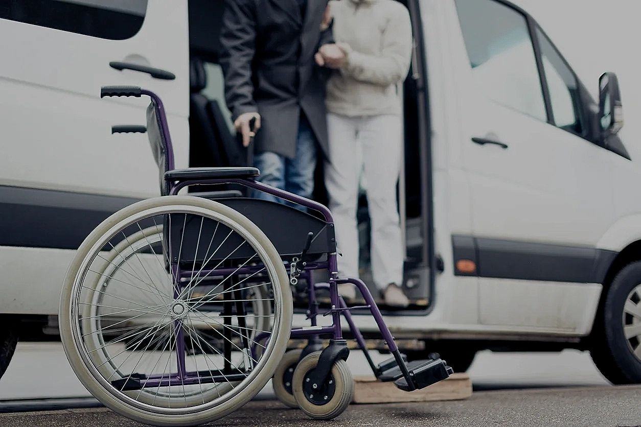 A wheelchair beside a van with a person assisting another out of the vehicle.