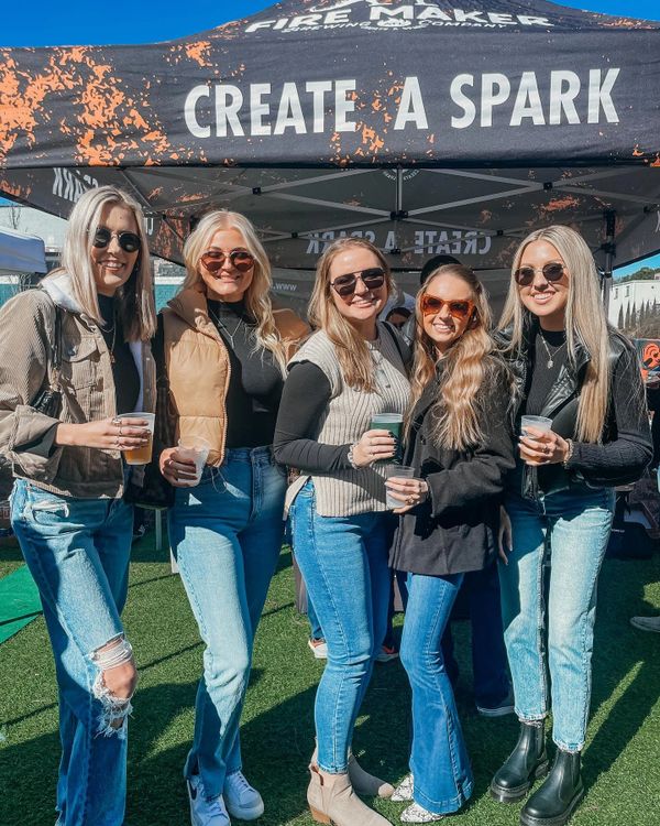Five women wearing sunglasses and casual outfits pose happily under a tent.