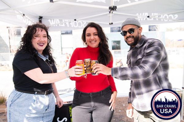 Three friends cheer with canned drinks under a tent at a bar crawl event.