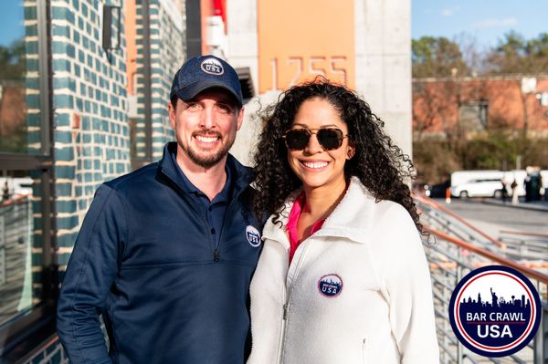 Two smiling people wearing Bar Crawl USA jackets outdoors.