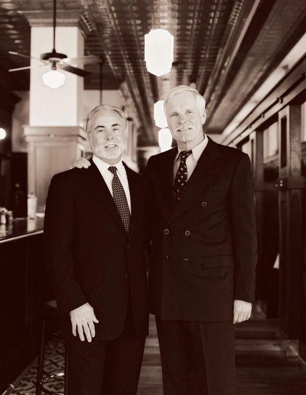 Two elderly men in suits posing in a vintage-style hallway.