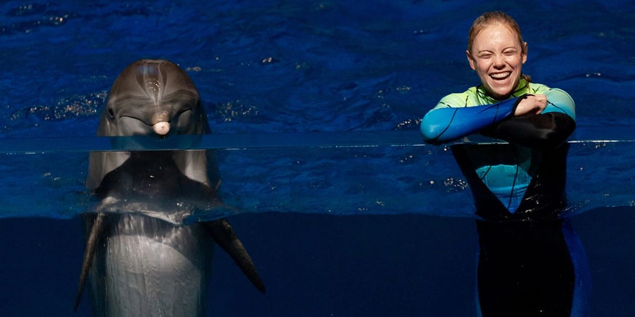 A happy woman in a wetsuit smiles next to a dolphin in the water.