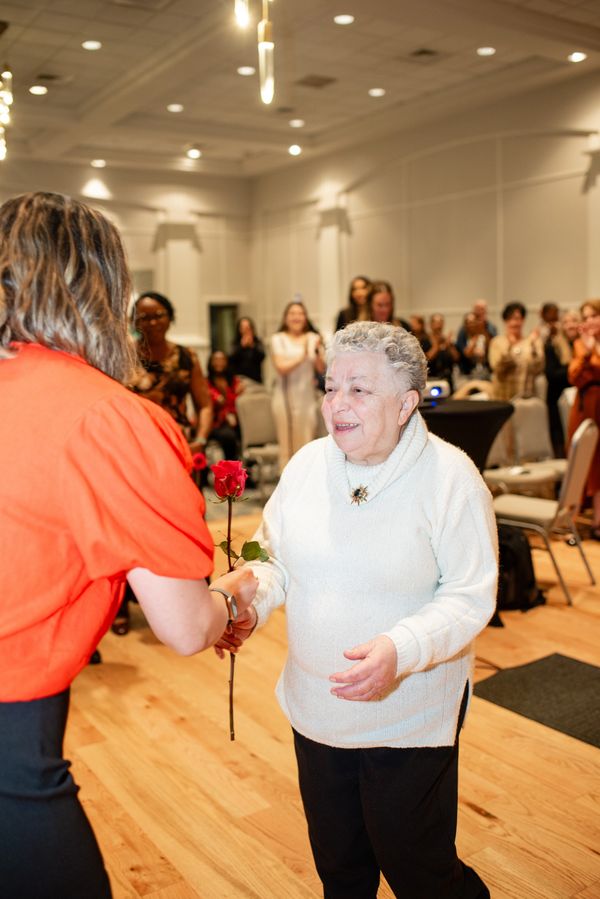 An elderly woman receives a red rose in a celebratory gathering.