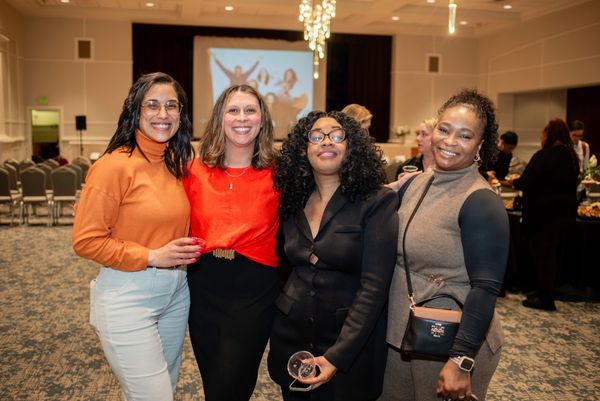 Four women smiling and posing at an indoor event with a projection screen behind them.