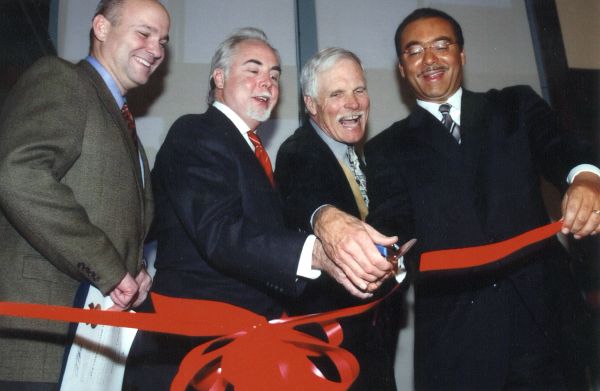 Four men in suits smiling while cutting a red ribbon at an event.