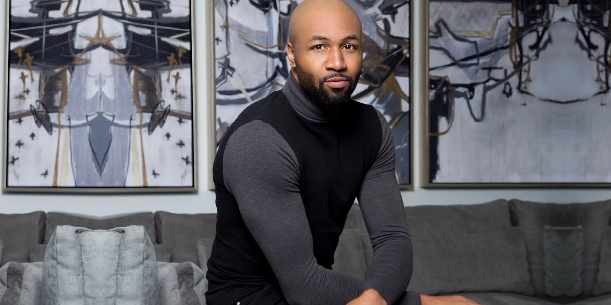 A man in a black and gray turtleneck sits on a gray couch with abstract art in the background.