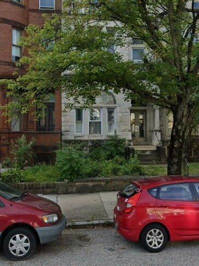 Two red cars parked by a sidewalk with trees and buildings.
