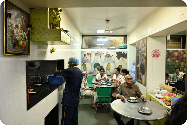 Man cooks in small kitchen as people enjoy meals, depicting a hospital canteen scene.