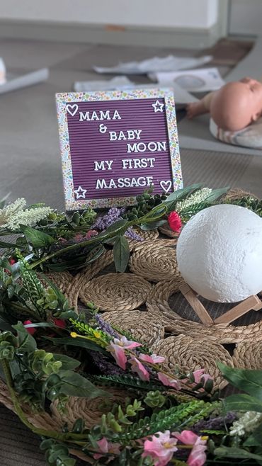 A sign announces a baby's first massage with decorative flowers and a moon prop nearby.