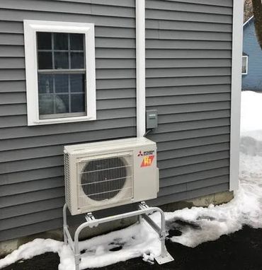 Outdoor air conditioning unit mounted on a gray house wall with snow on the ground.