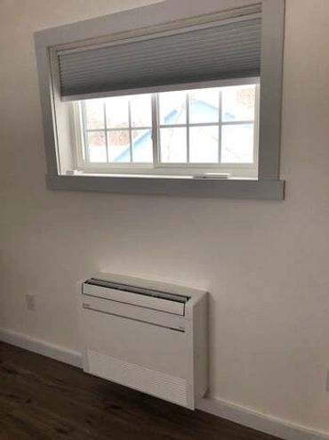 A wall-mounted air conditioner under a window with a partially closed blind.