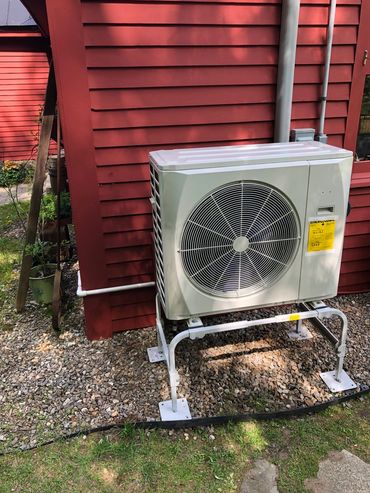 Outdoor air conditioning unit installed on a metal stand beside a red house.