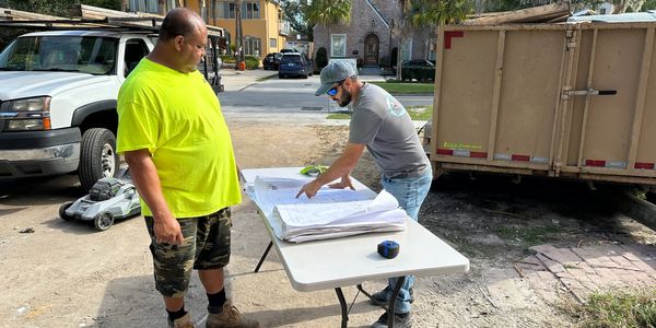 Two construction workers reviewing blueprints outdoors by a truck and trailer.