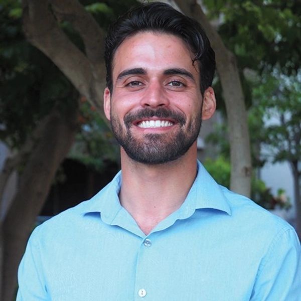 Smiling man in a light blue shirt outdoors with trees in the background.