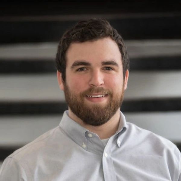 Smiling man with beard in a light shirt, casual office setting.