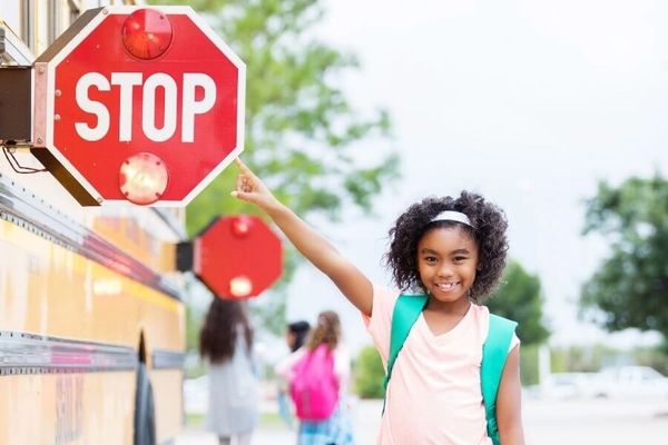 Smiling girl points to stop sign on school bus with other children in background.