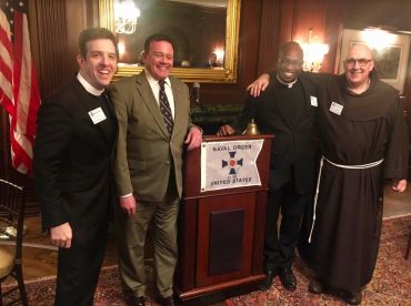 Four men in formal and religious attire posing by a podium with a Naval Order United States sign.