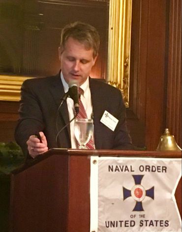 A man in a suit speaks at a Naval Order of the United States event.