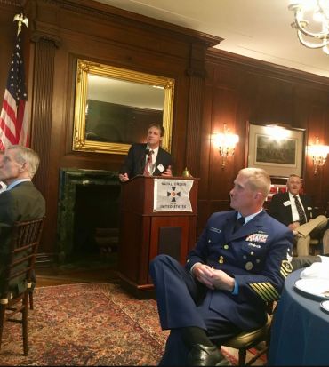 Man speaking at a podium in a formal room with military personnel seated.