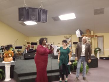 Three people stand near a stage, one speaking into a microphone, in a decorated indoor venue.