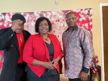 Three people posing happily in front of a festive backdrop with red and white lights and hearts.