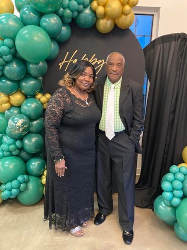 A couple dressed elegantly at a celebration with green and gold balloon decor.