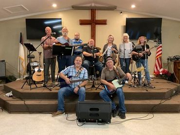 A church band poses on stage with instruments and a large wooden cross behind them.