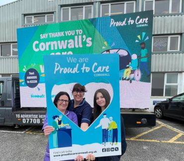 Three people pose with a 'Proud to Care Cornwall' frame in front of a promotional board.