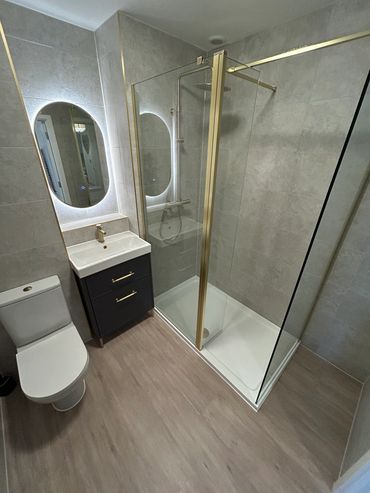 light grey wall tiles, white toilet, shower & basin with gold fittings and brown laminate floor