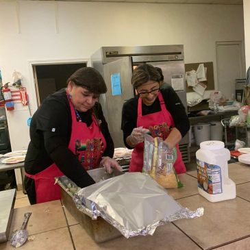 Two women preparing food in a kitchen wearing aprons.