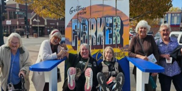 Group of elderly women sitting and smiling in front of a colorful Parker, Colorado sign.