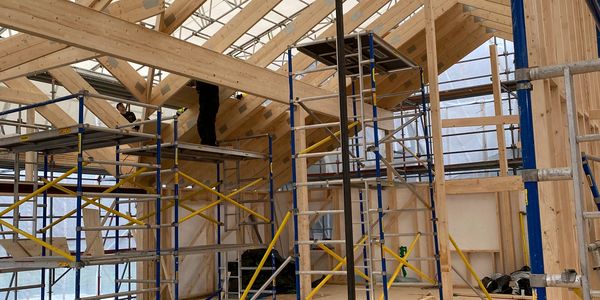 Wooden house frame under construction with scaffolding inside a temporary shelter.