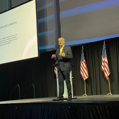 Speaker presenting on stage with American flags behind him.
