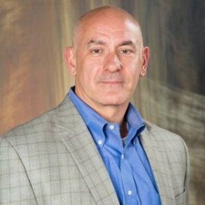 Professional portrait of a bald man in a blue shirt and checked jacket.